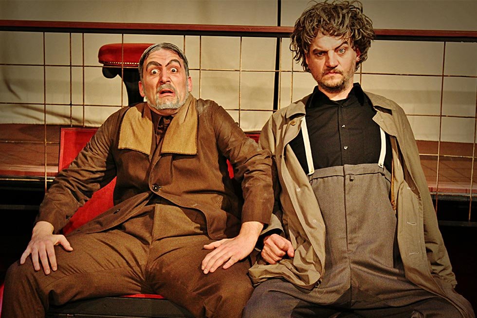 Deux acteurs dans Les Rustres de Goldoni : Thibault Moës avec une expression de surprise exagérée et un homme à l'air renfrogné, sur scène.